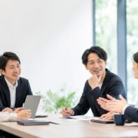 32e2d96e-248d-46d8-a7bc-1ce5d88a6ce7 | 一般社団法人日本セルフエスティーム普及協会|JISE|自己肯定感の専門団体 Five professionals seated around a conference table, chatting and smiling during a team meeting in a bright office.