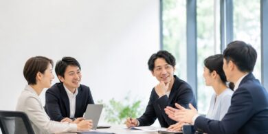 Five professionals seated around a conference table, chatting and smiling during a team meeting in a bright office.