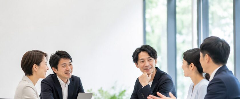 Five professionals seated around a conference table, chatting and smiling during a team meeting in a bright office.
