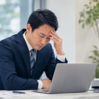db4555c8-a2e6-4437-8015-7acc41ed0be6 | 一般社団法人日本セルフエスティーム普及協会|JISE|自己肯定感の専門団体 Man in a navy suit sitting at a desk with a laptop, head resting on his hand in a stressed pose.