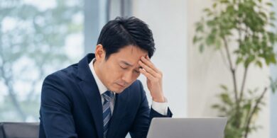 Man in a navy suit sitting at a desk with a laptop, head resting on his hand in a stressed pose.